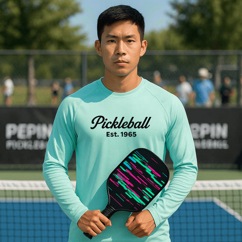 Person wearing a turquoise 'Pickleball Est. 1965' shirt holding a pickleball paddle on a court.